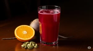 Um copo de vidro com kvass de beterraba de cor vermelha vibrante, decorado com uma fatia de laranja e sementes de cardamomo ao lado, sobre uma mesa escura.